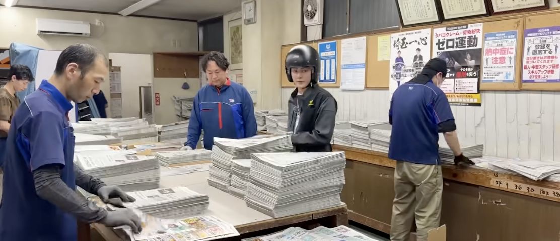 新聞販売店でスタッフと新聞を準備する三崎優太